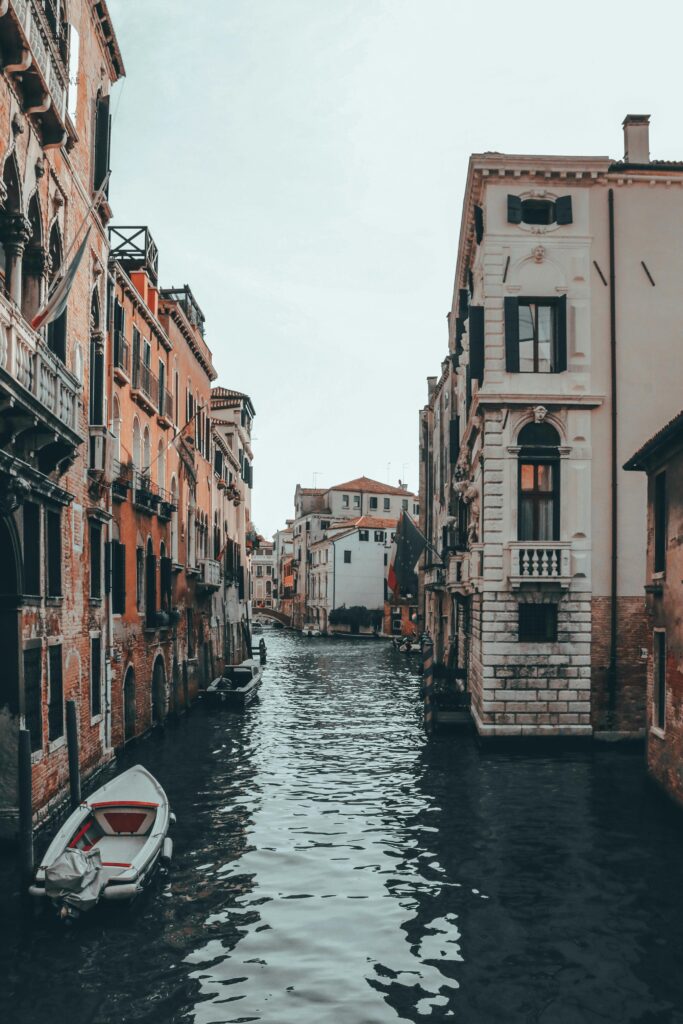 Boats on canal with ripples between old multistory stone building facades under light sky in Venice